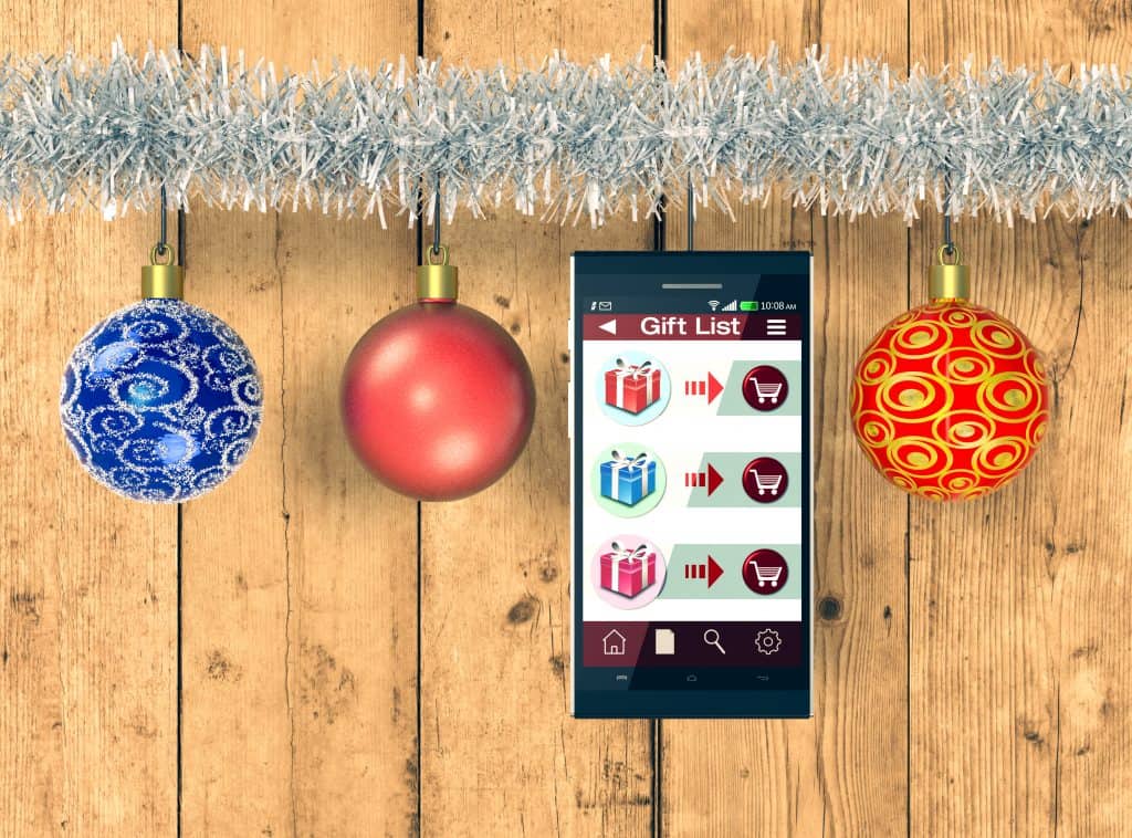 A phone with "Gift List" on the screen hanging from a tinsel garland surrounded by ornaments