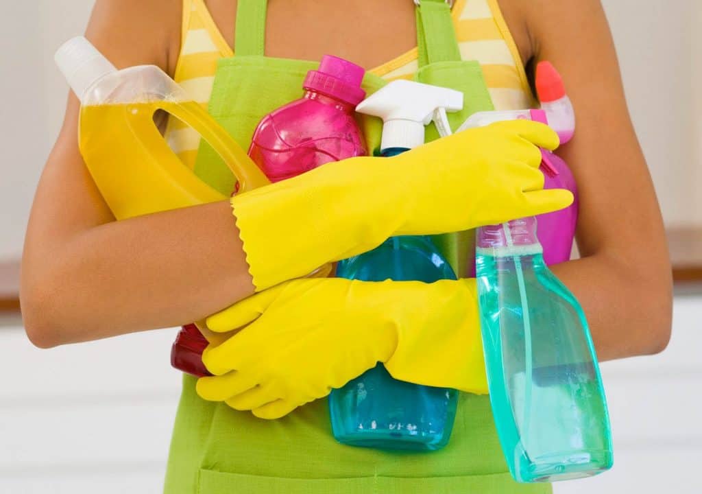 Closeup of torso of woman in apron wearing yellow rubber gloves and holding cleaning products.