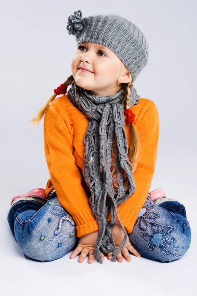 A young girl in a gray knit cap and scarf, orange shirt and blue jeans.
