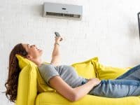 Young woman lying on yellow couch with remote control enjoying the breeze from an air conditioning unit
