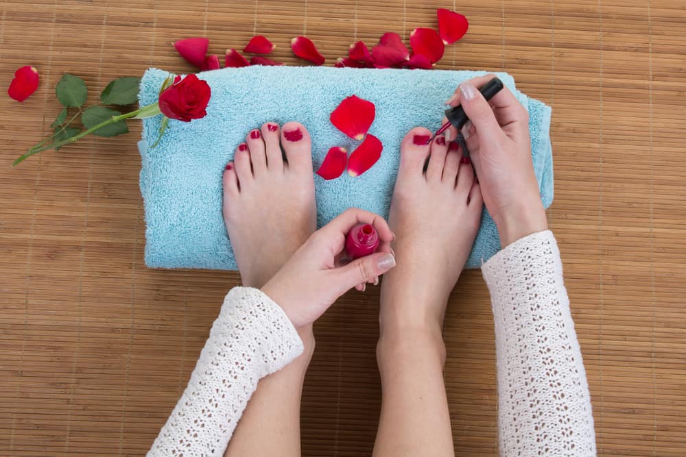 A woman painting her toenails red, with feet resting on a blue towel surrounded by rose petals.
