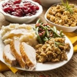 A plate with turkey, green beans and dressing, surrounded by a bowl of cranberry sauce and other traditional Thanksgiving side dishes.