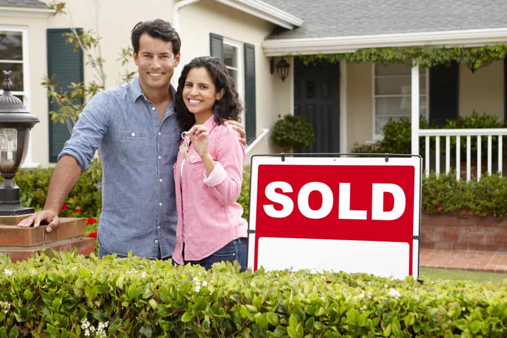 A male and female stand near a "Sold" sign in front of a house. The woman is showing thumbs-up.