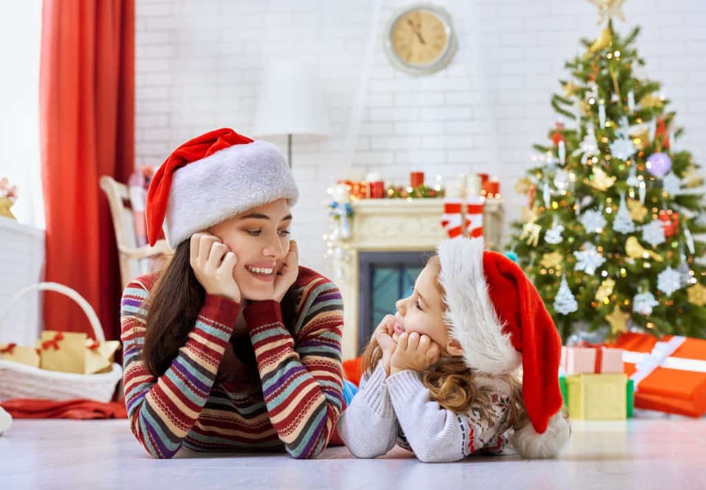A mom and daughter in Santa hats in a living room decorated for Christmas, lying on the floor looking at each other lovingly.