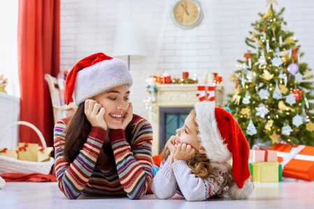 A mother and young daughter in Santa hats lie on the floor looking at each other lovingly.