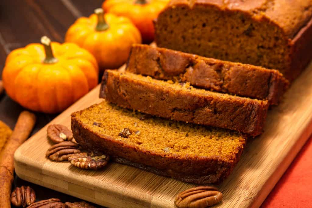 Sliced pumpkin bread on wooden cutting board surrrounded by small pumpkins.