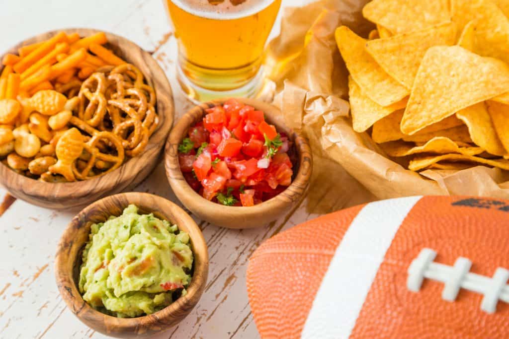 Football party food -- chips, salsa, guacamole, pretzels and beer -- next to a football on a table.