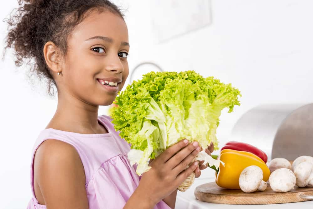 African American girl holding leaf lettuce next to a table with peppers and mushrooms.