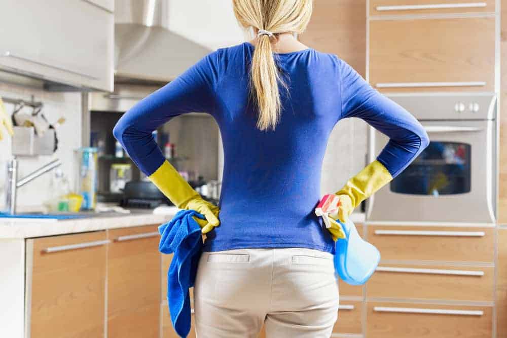 Woman in rubber gloves holding cleaning supplies in kitchen.