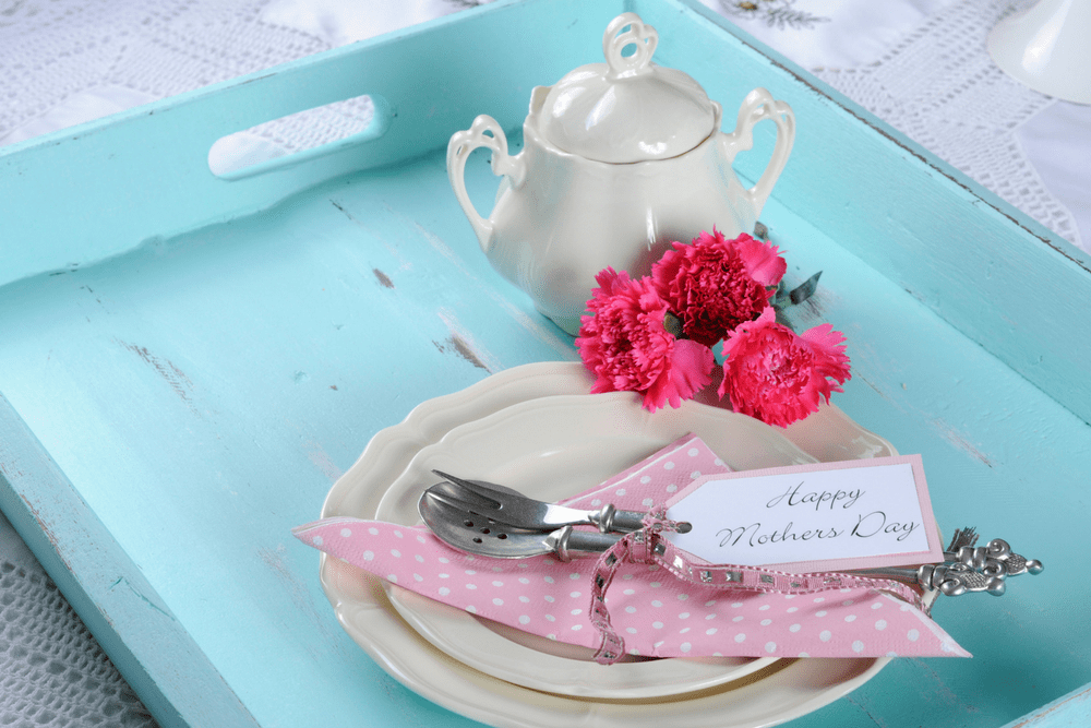 A turquoise wooden tray with a white teapot, pink flowers, white china plate and pink napkin with a card that says "Happy Mother's Day."