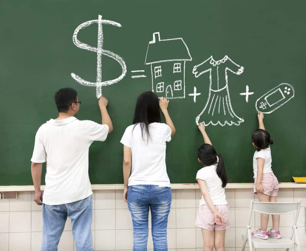 Family of mom, dad and 2 children at green chalkboard drawing dollar sign, house, clothes and remote control.