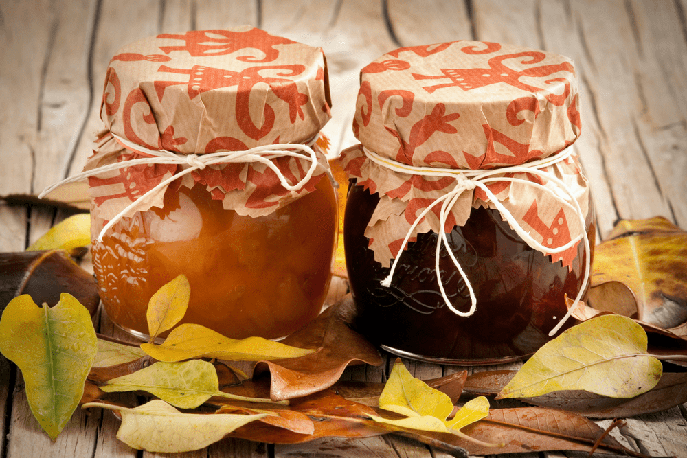 Small jars of jam with paper covers tied with raffia, on a wooden board surrounded by leaves.