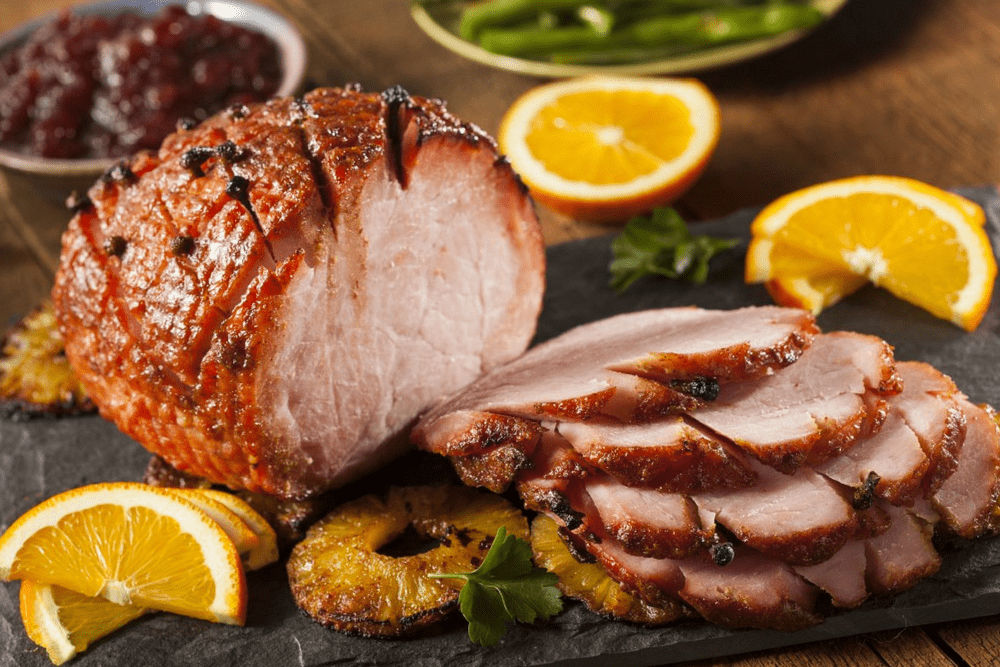 Easter ham surrounded by orange slices with green beans and cranberry sauce in the background.