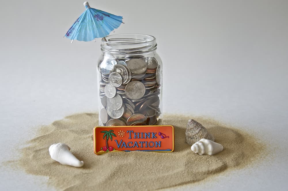 A jar of coins on a pile of sand with seashells and a paper umbrella tucked in it, with a sign saying "Think Vacation."