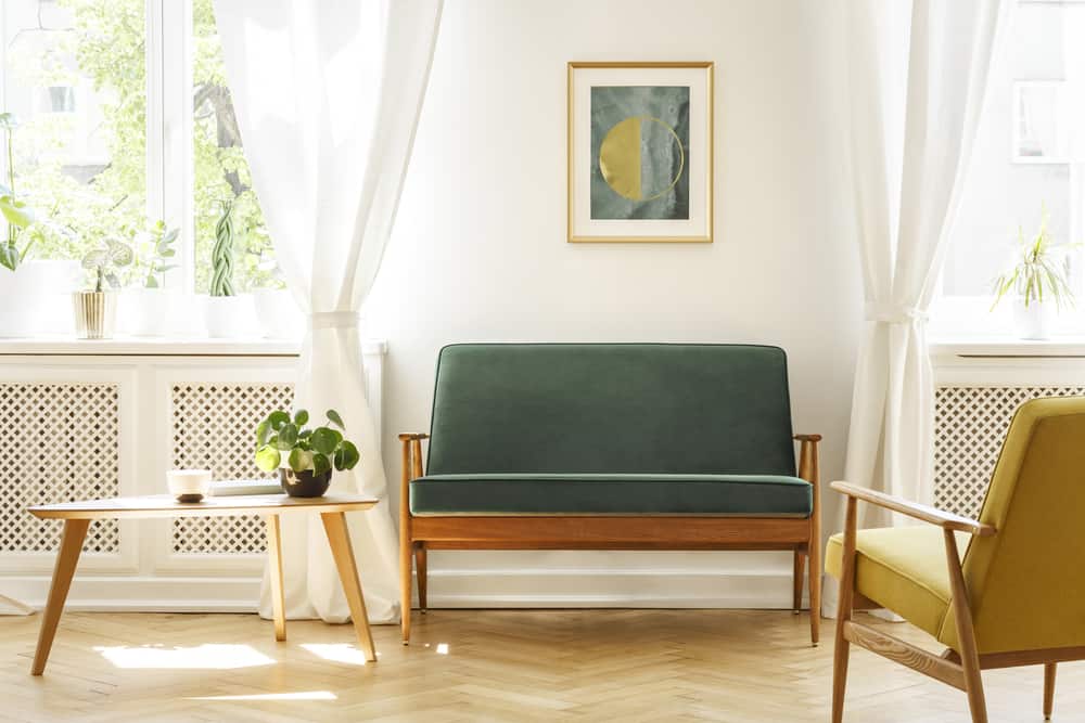 A green midcentury modern settee against a white wall with windows behind it.