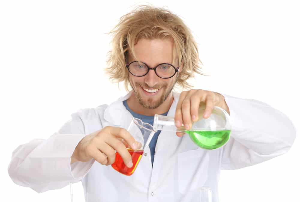 Blond man with wild hair and glasses wearing lab coat and pouring a beaker of green liquid into a beaker of red liquid.