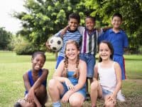 6 boys and girls of diverse races and ethnicities kneel or stand for a photo in a park. One boy holds a soccer ball.