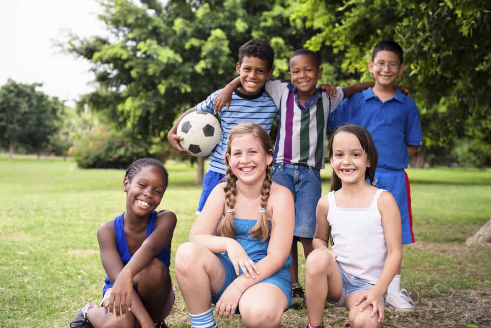 6 boys and girls of diverse races and ethnicities kneel or stand for a photo in a park. One boy holds a soccer ball.