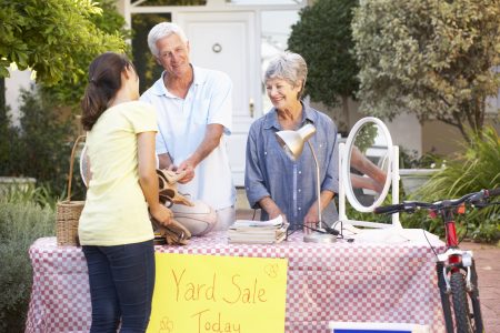 Make money with Older couple behind table with red-and-white checkered tablecloth and sign saying Yard Sale Today selling to young brunette woman in yellow