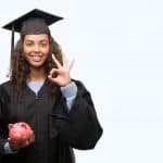 Money tips for graduates - Young hispanic woman wearing graduation uniform holding piggy bank doing ok sign with fingers, excellent symbol