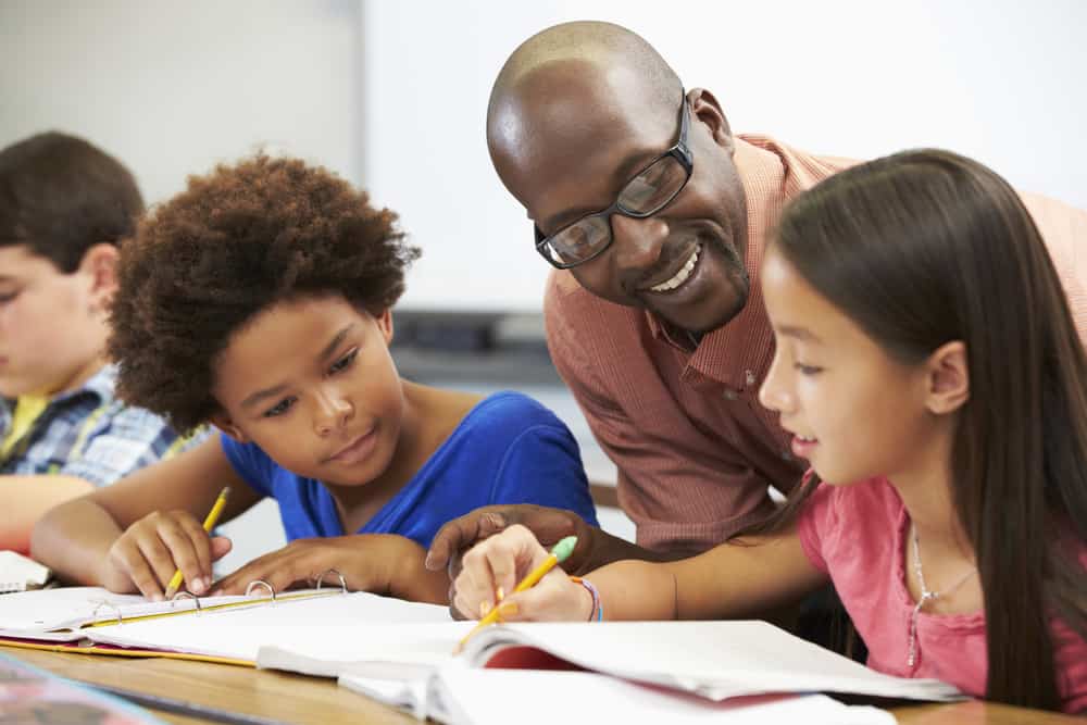 A Black male teacher leans over to help a Black child and a Latina child with their classwork.