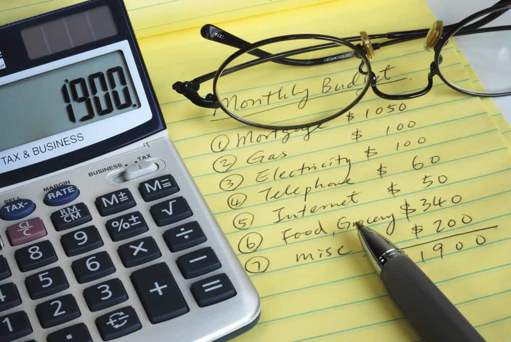 Calculator, glasses and pen on top of yellow legal pad with "Monthly Budget" and categories written on it.