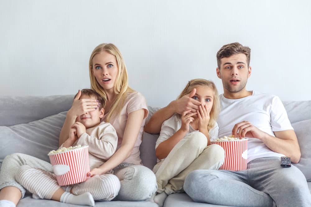 Young couple putting their hands over children's eyes while sitting on a couch with popcorn.