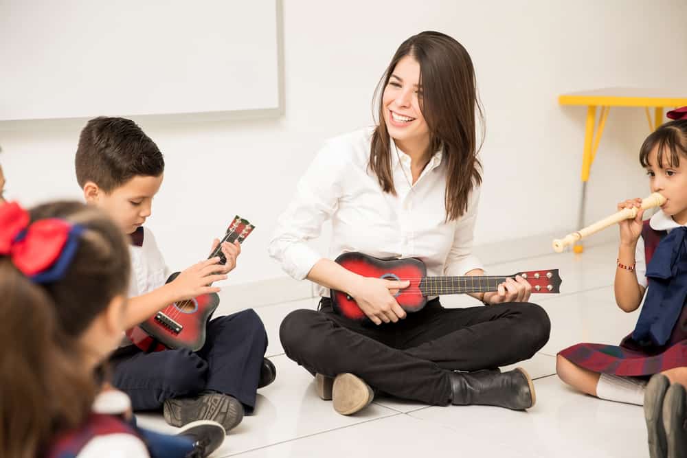 A woman seated on the floor plays the ukulele surrounded by children holding other instruments.