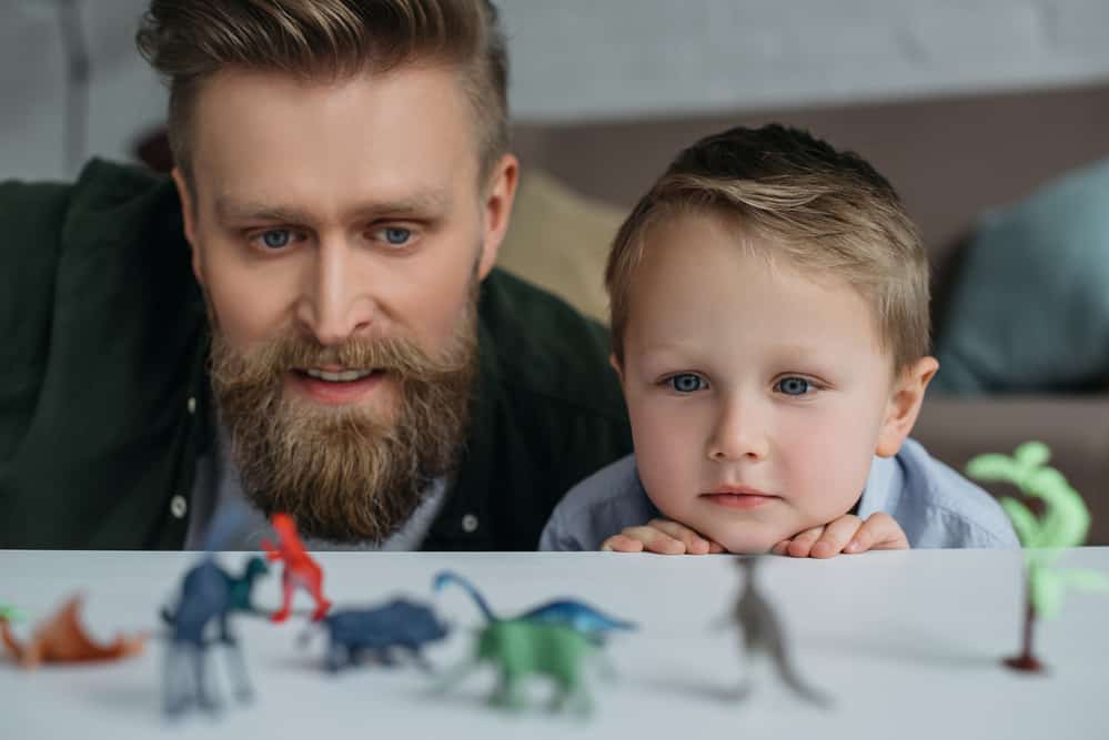 A father and his son look at toy dinosaurs on a table.