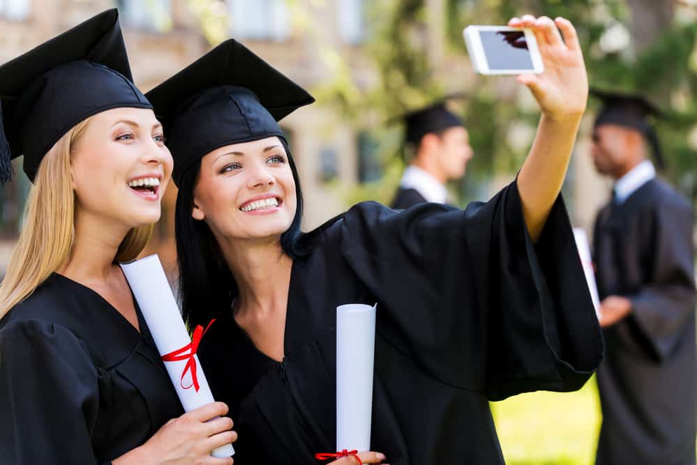 Two young women taking a selfie in graduation gowns with diplomas.