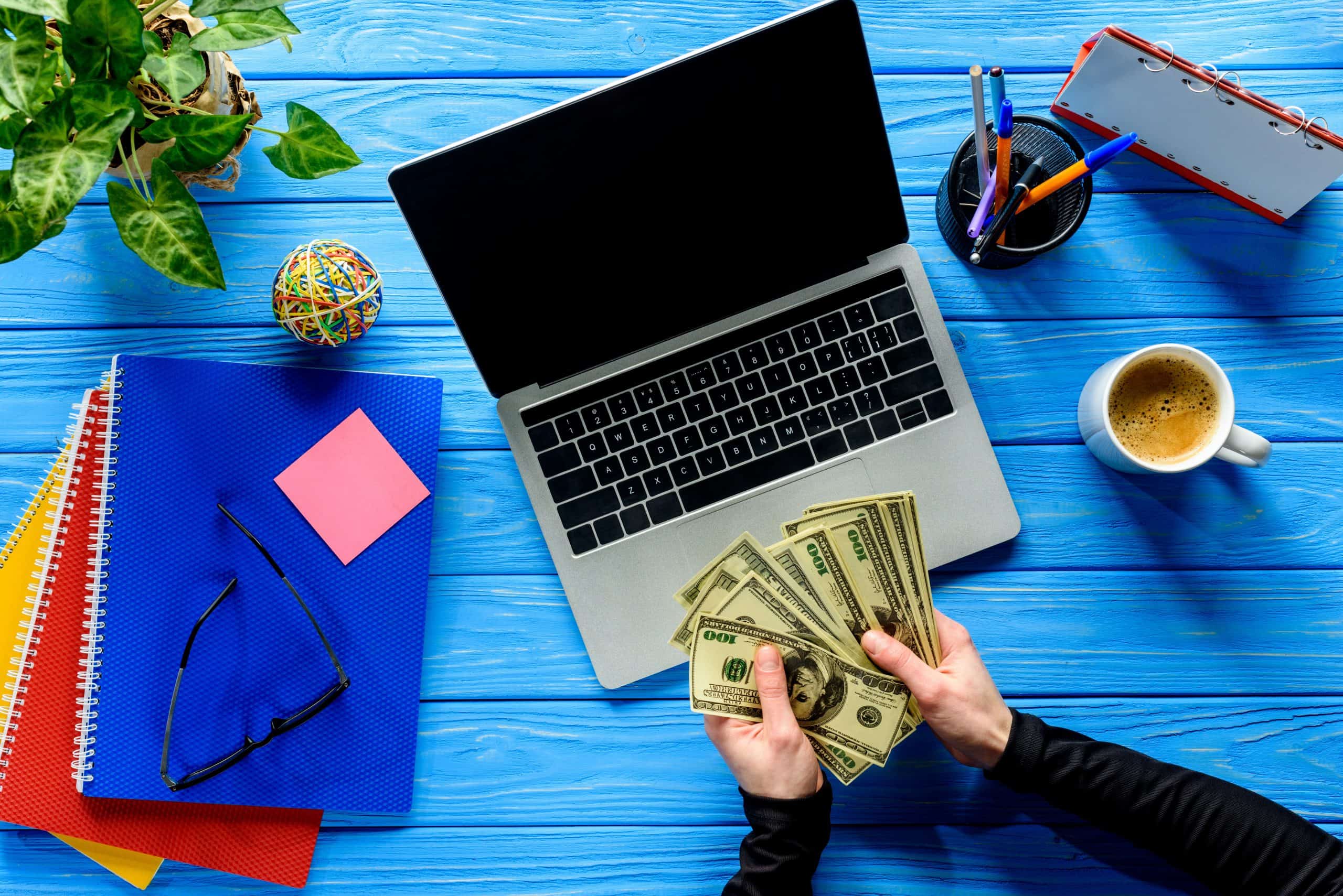 Spend money to save time - hands holding hundred dollar bills by laptop on blue desk