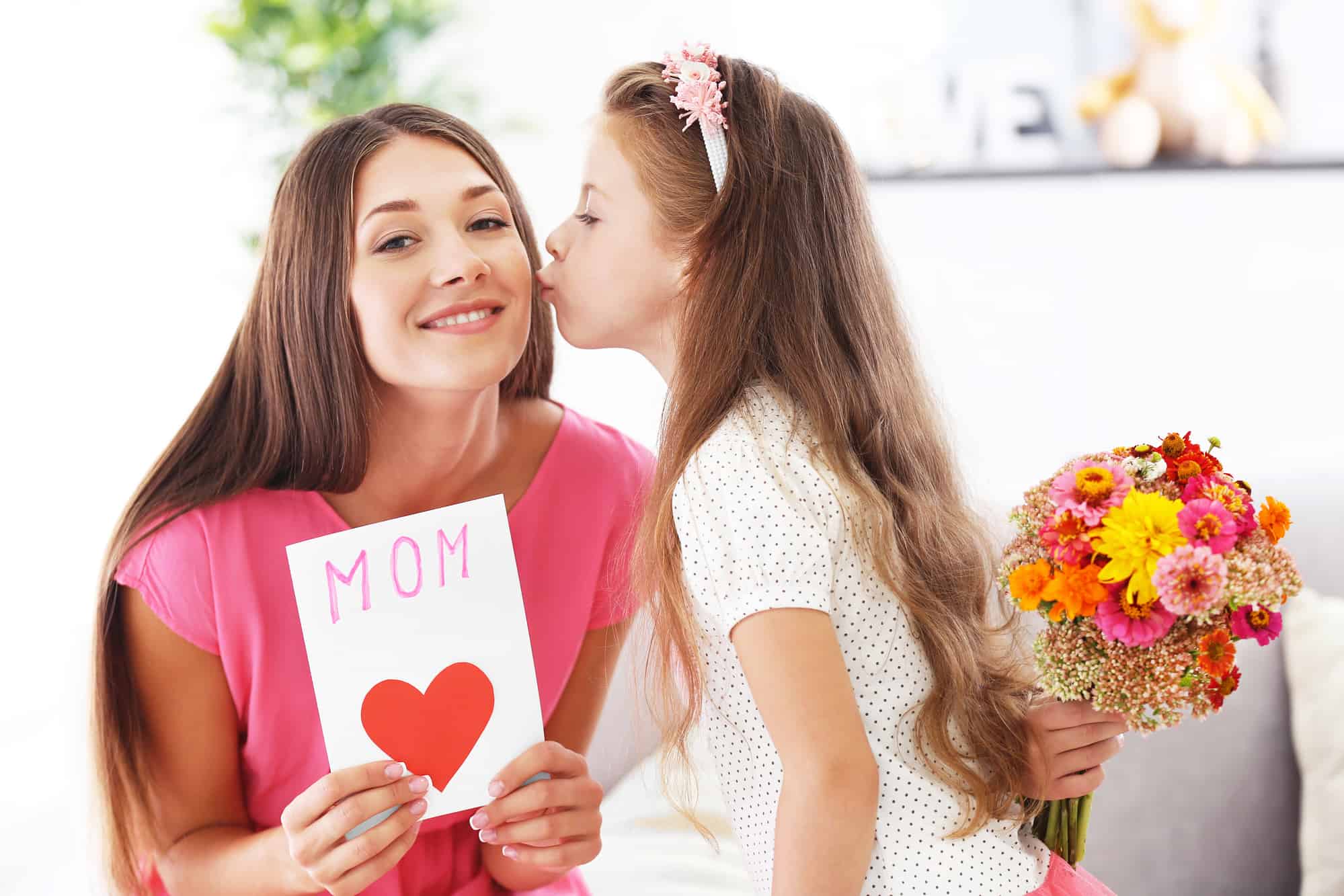 A young mother holding up a handmade Mother's Day card and getting a kiss from her daughtr