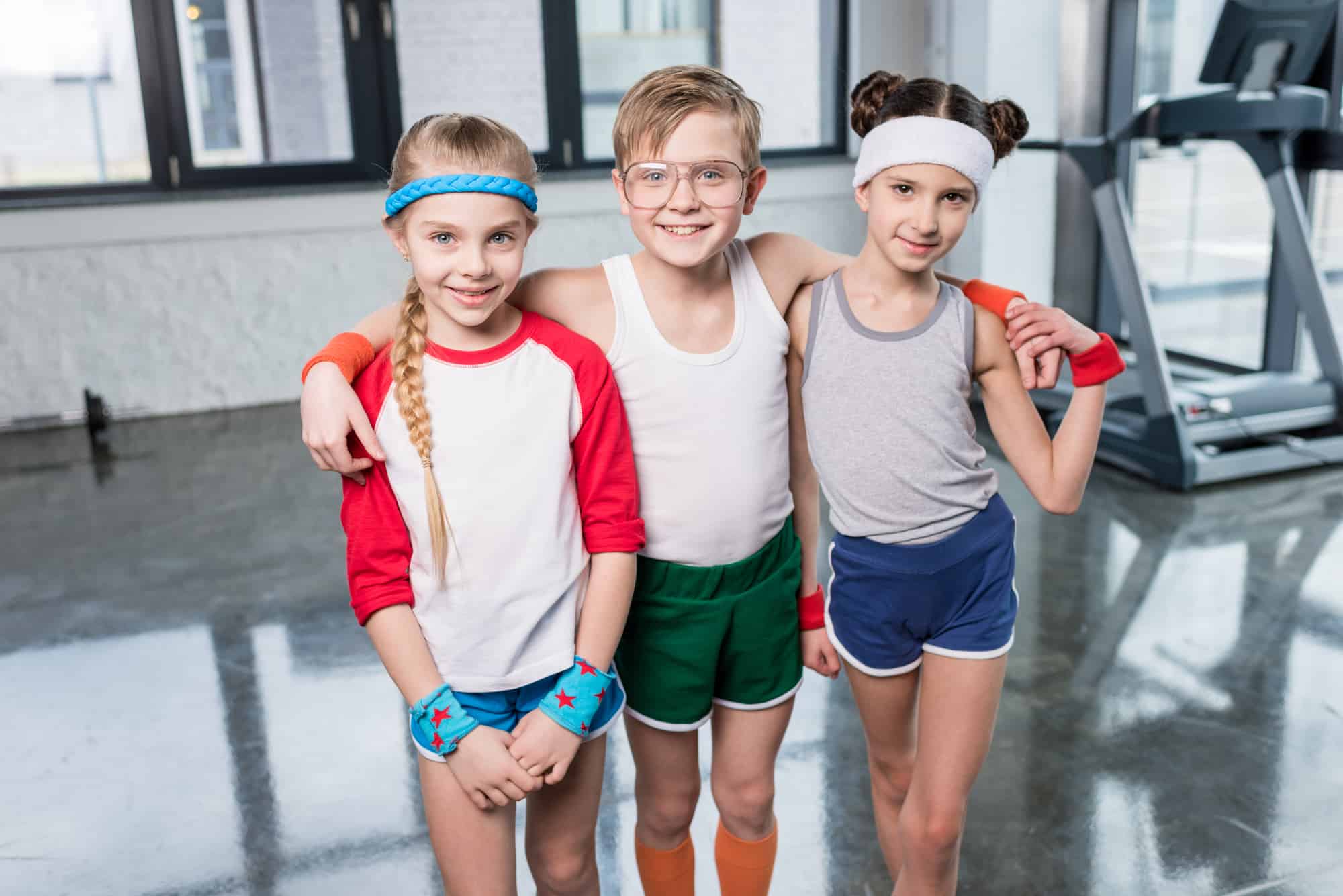 A young white boy with his arms around the shoulders of two white girls. They are in a gym dressed in shorts and cotton shirts.