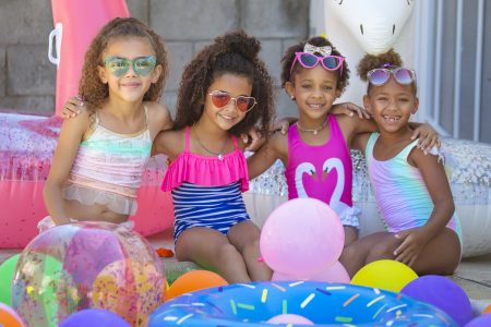 4 young girls of color in bathing suits surrounded by balloons and pool toys at a pool party.