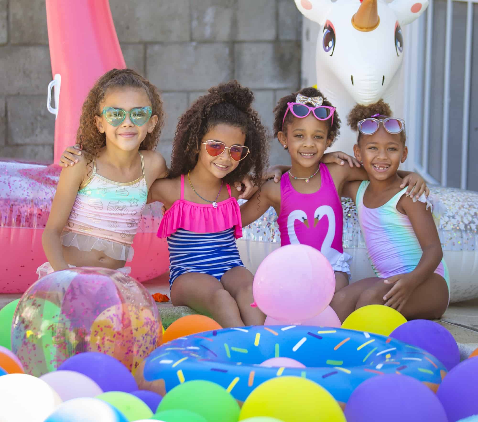 4 young girls of color in bathing suits surrounded by balloons and pool toys at a pool party.