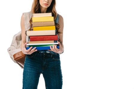 A young white woman in jeans carries a stack of textbooks.