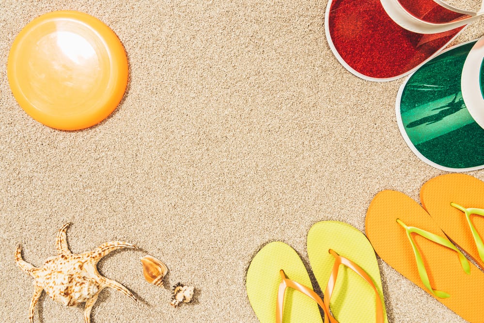 From top right, two sun visors and two pairs of flip flops on a sandy beach next to a crab and a plastic Frisbee.