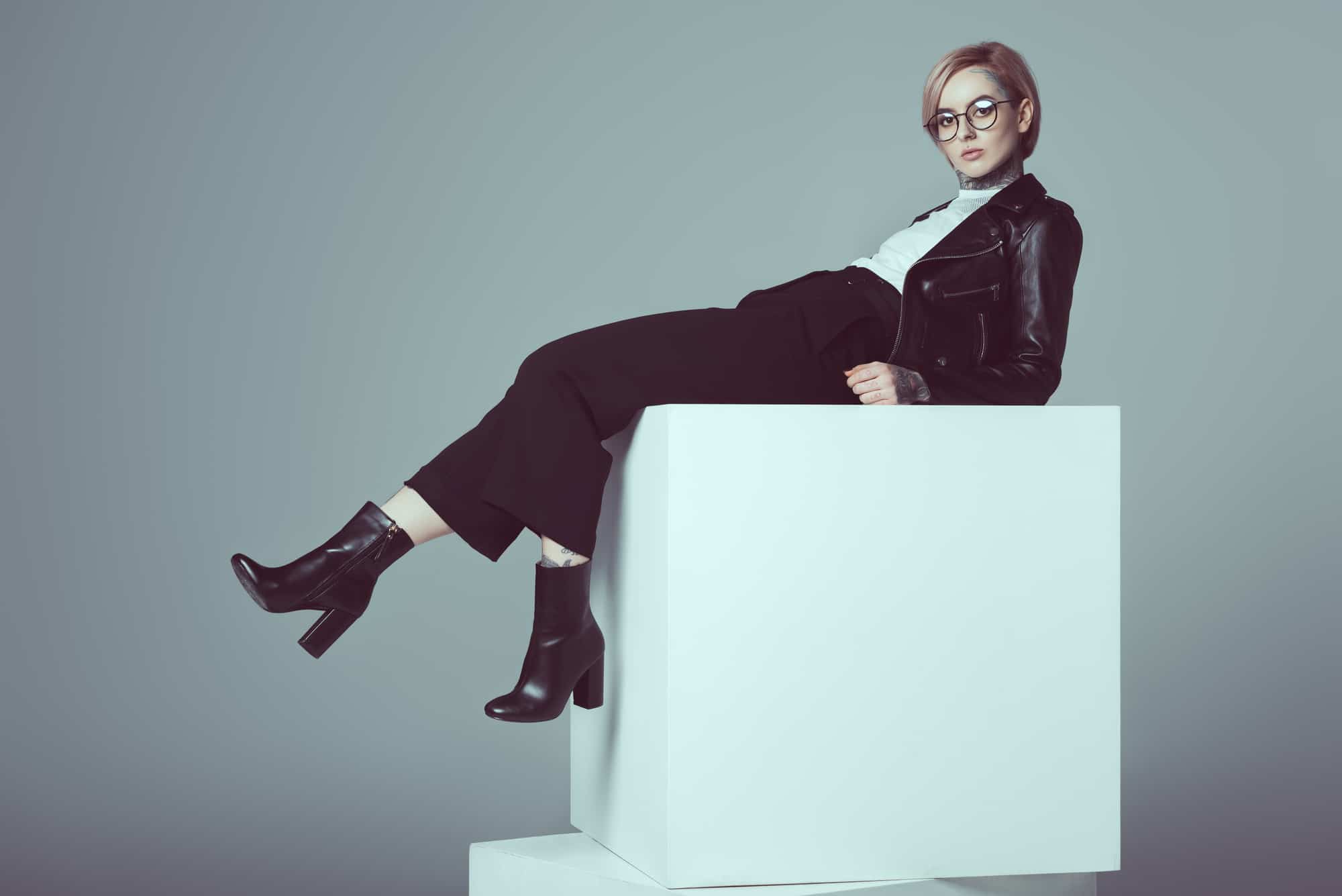 Blond woman in black pants, black boots, black leather jacket and white blouse leaning back on large white block.