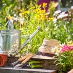 A straw hat, tin watering can and gardening tools on steps in a garden surrounded by flowers.