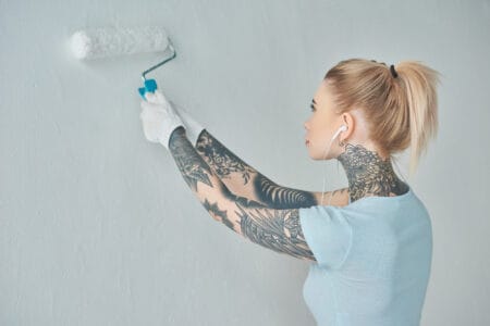 Young woman paints a wall a muted gray while wearing earbuds and listening to music.