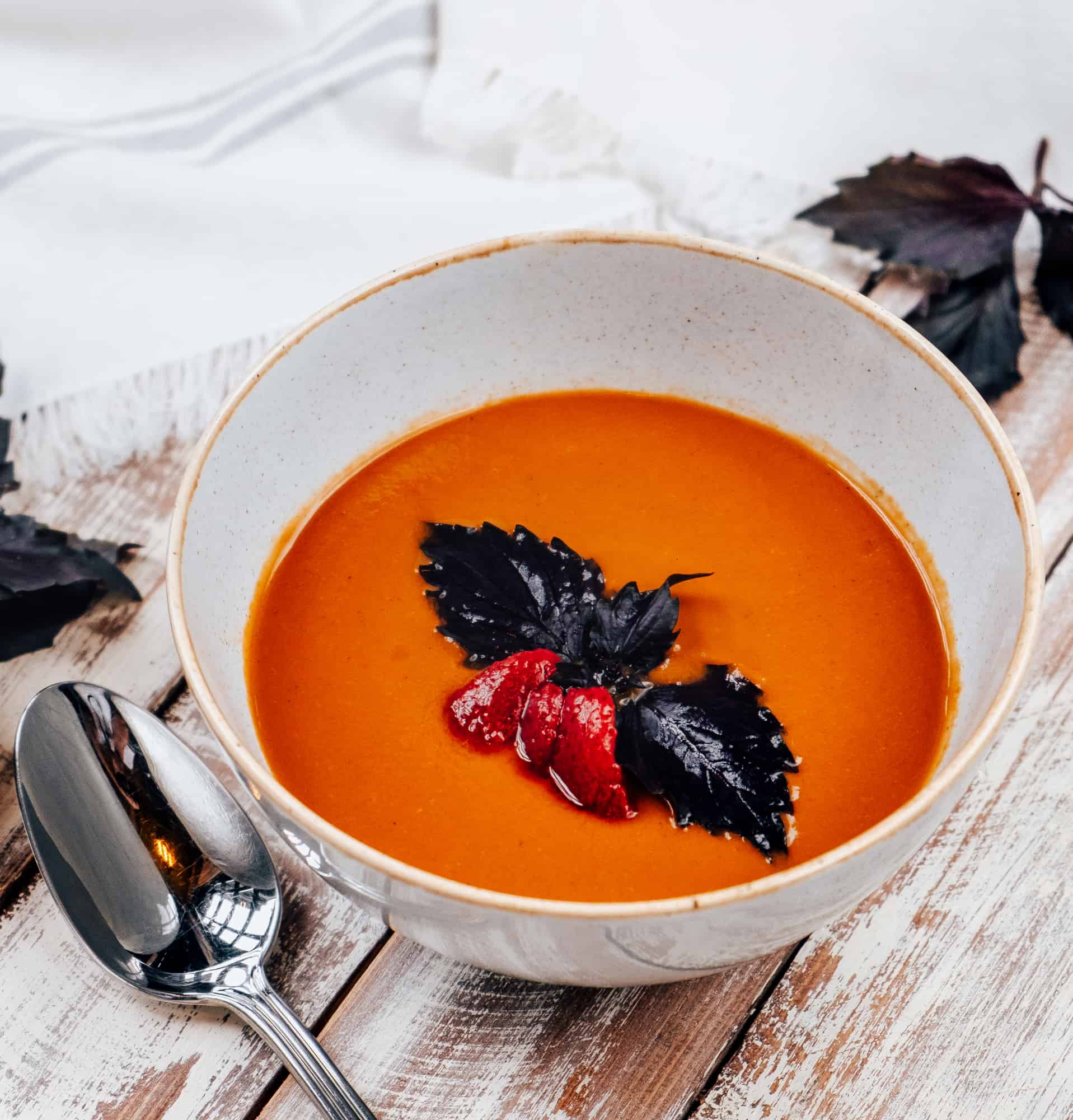 Tomato soup garnished with purple basil in a white bowl on a wooden table with a spoon at the side.