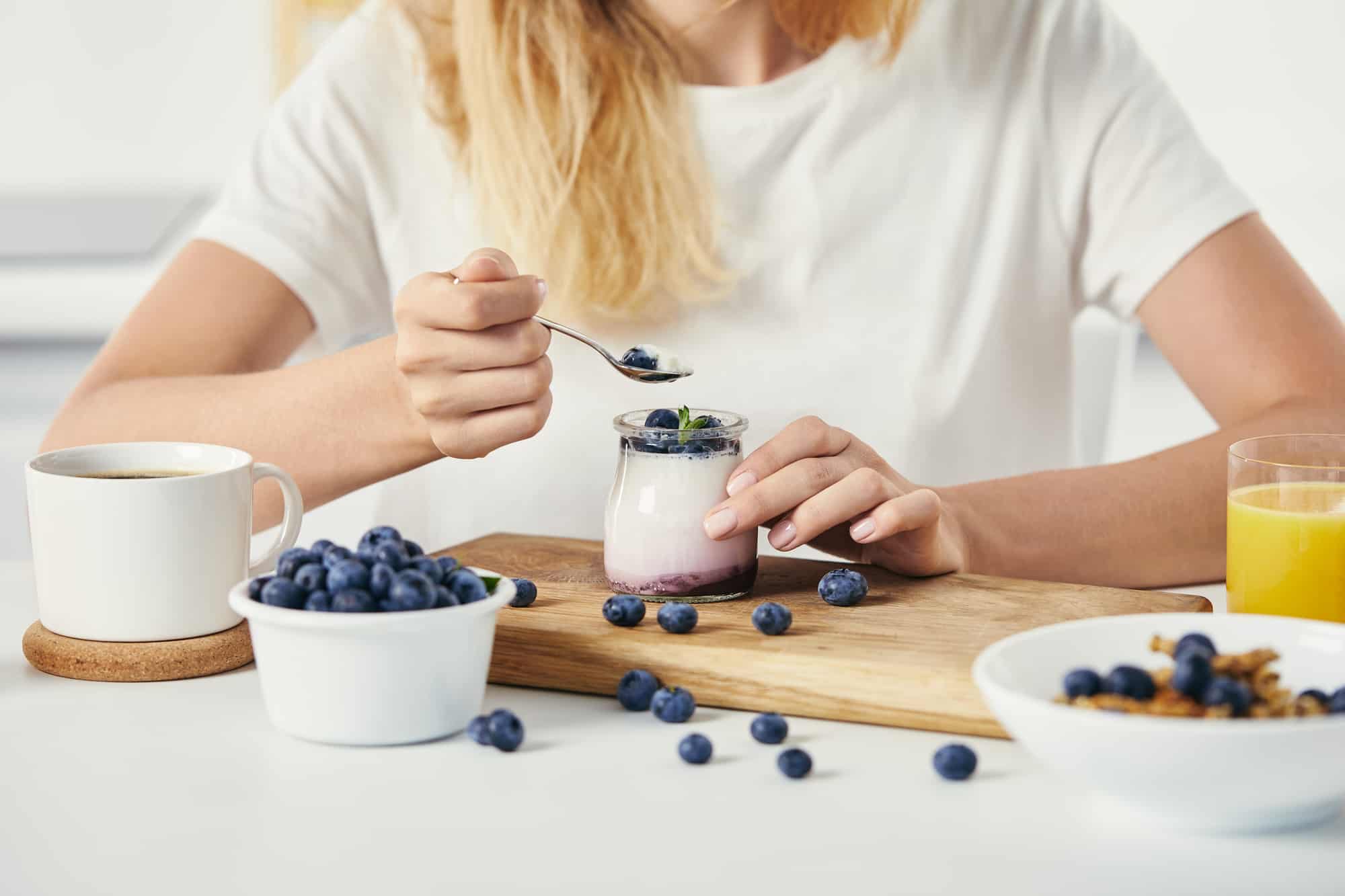 Blonde woman in white t-shirt eating blueberries and yogurt from a glass jar with coffee and orange juice on table