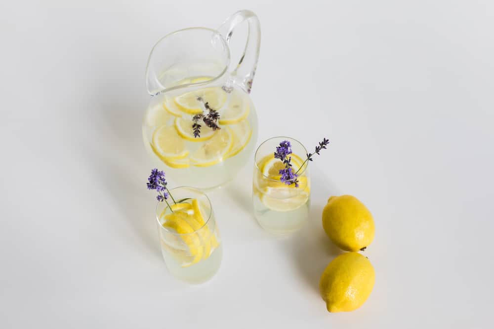 A pitcher of water with sliced lemons, two glasses with lavender sprigs in them alongside two whole lemons.