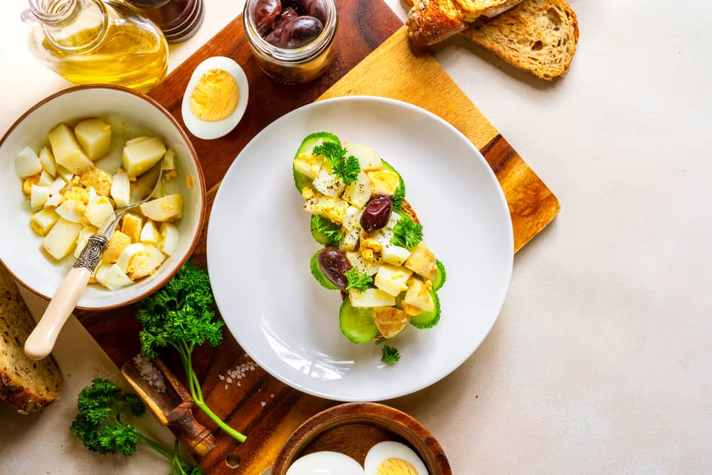 A cutting board with a bowl of potatoes, a hard boiled egg, some parsley and potato salad on a plate.