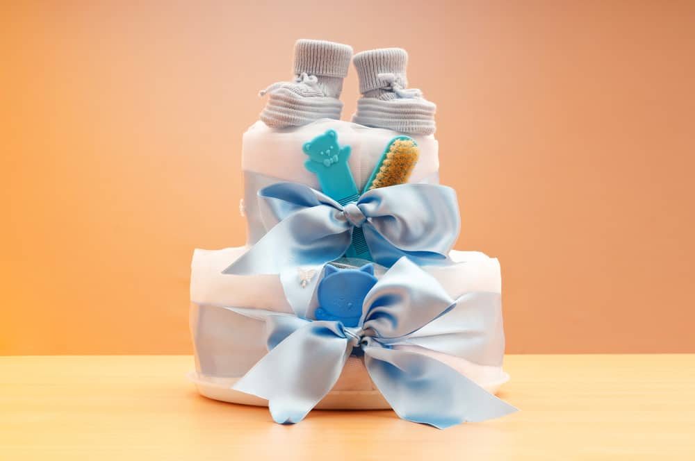 Un pastel de dos pisos hecho de pañales apilados atados con una cinta azul con botitas de bebé encima.  bebé 
