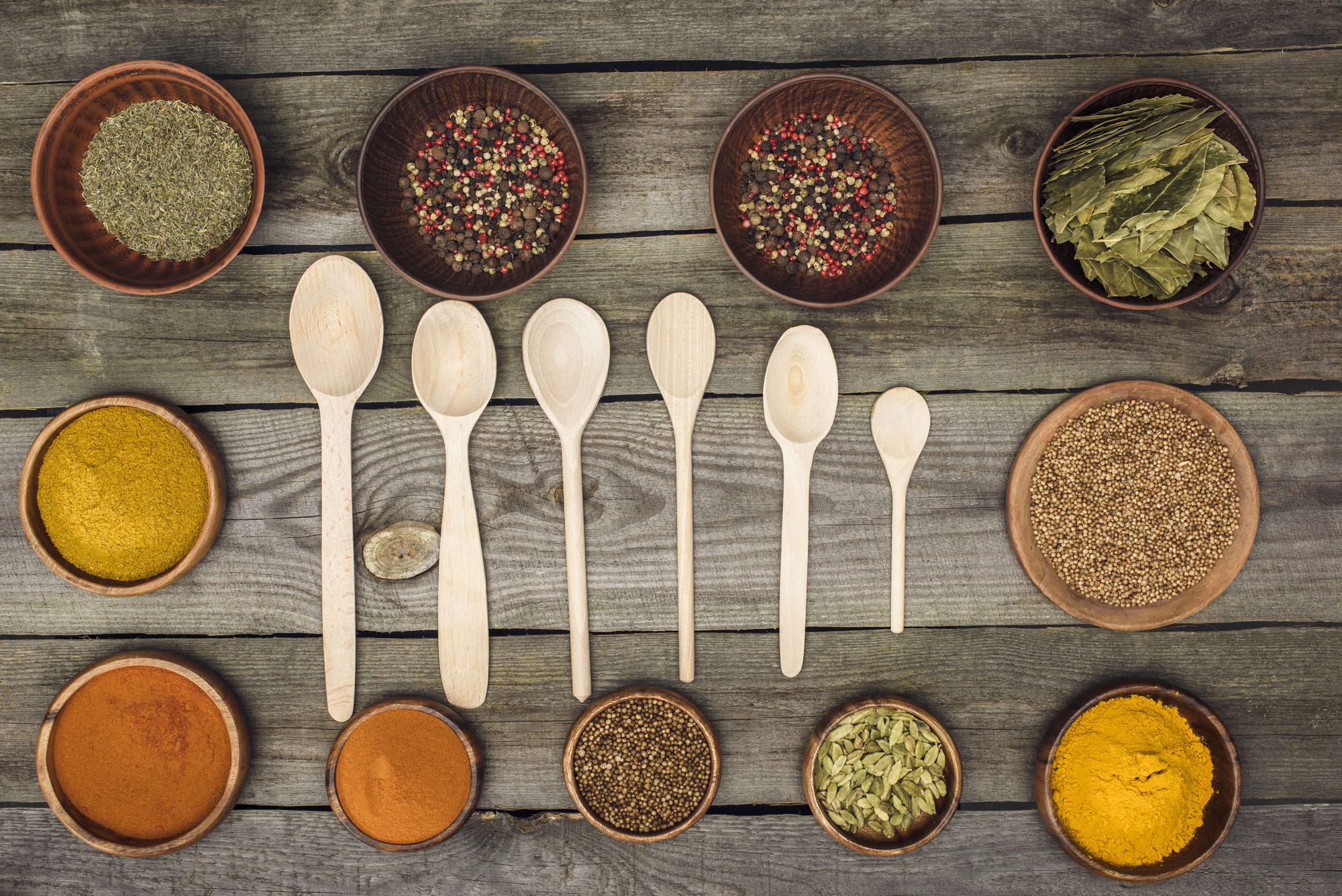 Wooden spoons surrounded by bowls of spices on a wooden board.