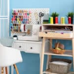 Sewing space with sewing machine on white desk with white pegboard and colorful spools of thread behind it.