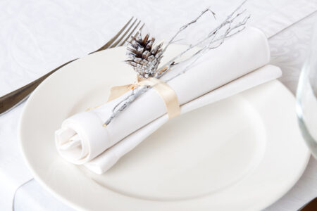 An all-white place setting with white plate and white napkin on white tablecloth, with a silver pine cone.