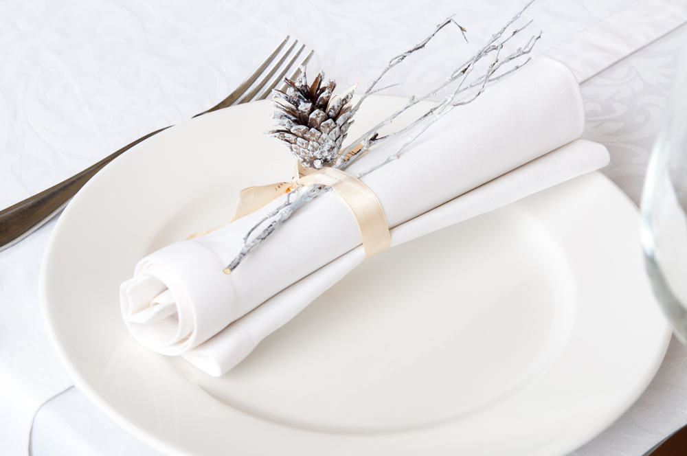 An all-white place setting with white plate and white napkin on white tablecloth, with a silver pine cone.