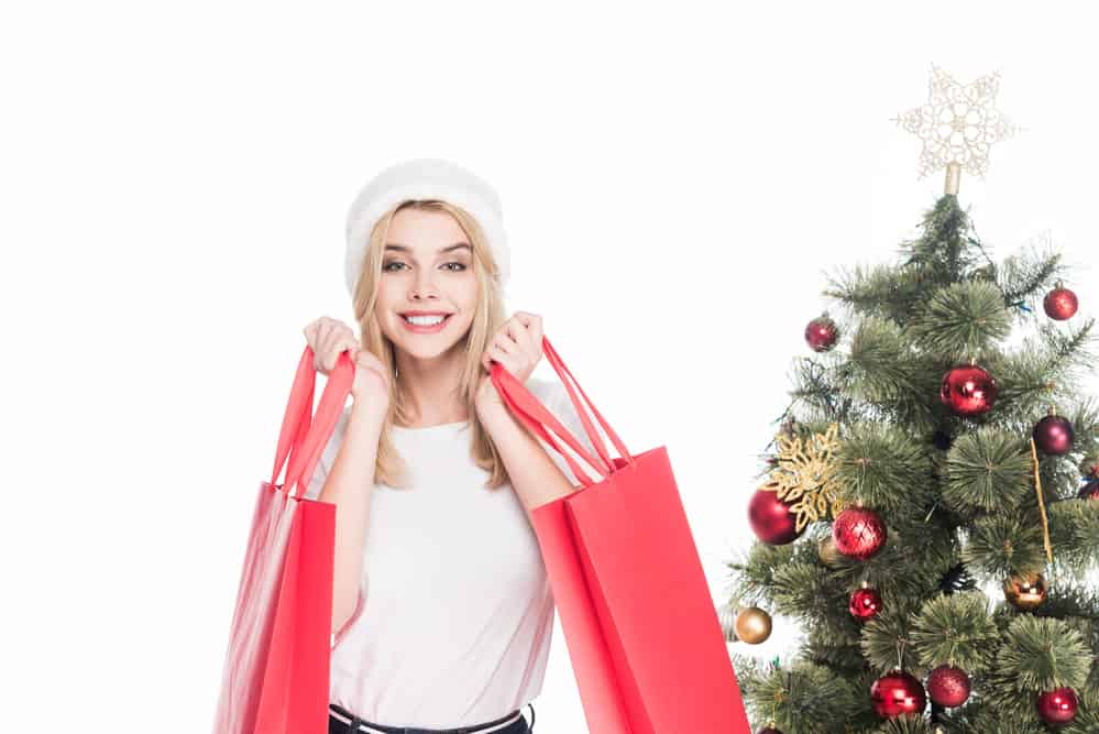 Woman wearing Santa hat carrying 2 shopping bags with Christmas tree in background